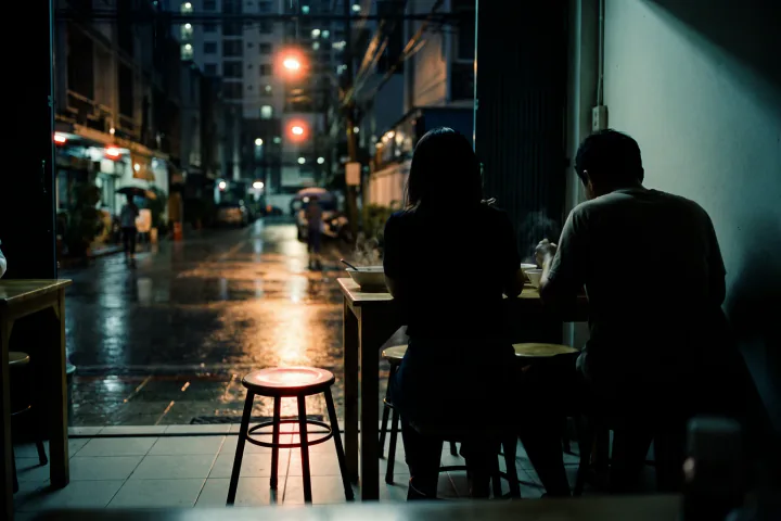 ร้านอาหารห้องแถวยามค่ำ คนสองคนนั่งกินข้าวด้วยกันที่โต๊ะเล็ก มองเห็นแค่ด้านหลังและด้านข้าง เก้าอี้ตัวที่สามถูกเลื่อนออกมาเล็กน้อย ว่างเปล่า มีแสงสีชมพูซีดตกกระทบ