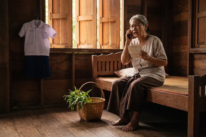 หญิงชรานั่งริมเตียงในบ้านไม้อีสานยามสาย มือหนึ่งแนบโทรศัพท์ไว้ที่หู อีกมือกำสมุดบัญชีธนาคารแน่น หน้าตึงเครียด
