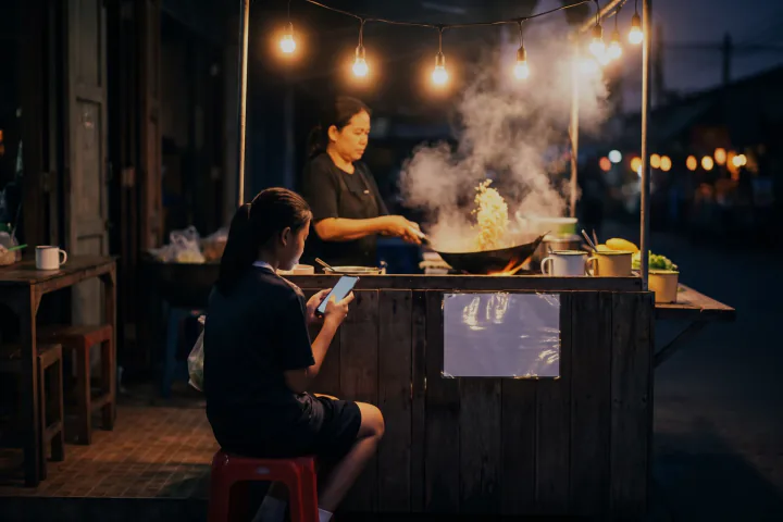 แผงข้าวผัดในตลาดนัดกลางคืน เด็กสาวนั่งถือโทรศัพท์ที่มีแสงนวล แม่ยืนผัดข้าวอยู่ข้างหลังท่ามกลางไอร้อนและไฟหลอดสีส้ม