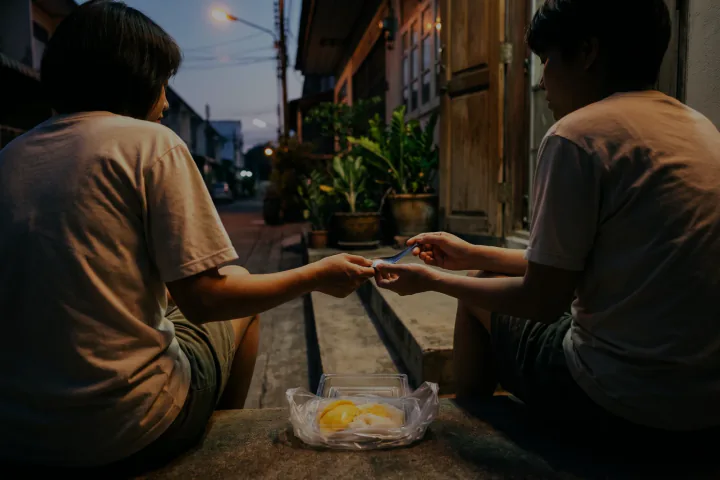 เพื่อนสองคนนั่งไหล่ชิดกันบนขั้นบันไดหน้าบ้าน คนหนึ่งยื่นช้อนให้อีกคน กล่องข้าวเหนียวมะม่วงเปิดวางอยู่ระหว่างทั้งคู่ ฟ้าปลายซอยเริ่มสาง
